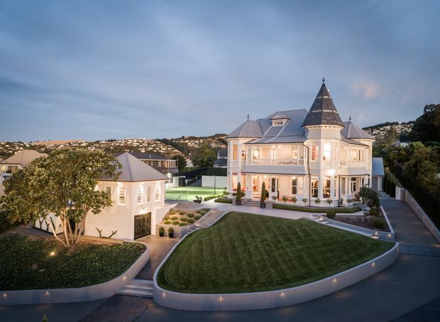 Aerial dusk view of a grand multi-storey residential villa with ornate white façades, multiple steep pitched roofs, conical turret roofs, balconies, and tall windows. The property includes a manicured lawn, curved landscaped garden beds, paved paths, a detached outbuilding, and surrounding suburban hills.