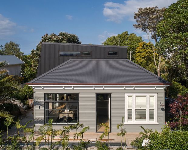 Front exterior view of a compact grey weatherboard house with a steep corrugated metal roof and a dark, box-like upper addition. The facade combines traditional horizontal cladding, a large black-framed multi-pane window, a narrow glazed entry door, and white louvered shutters, set within a lush landscaped garden.