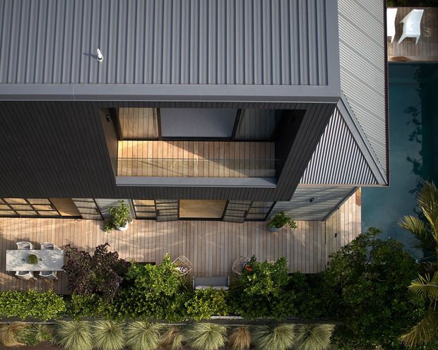 Top-down aerial view of a contemporary residential building with sharply articulated geometric forms, extensive dark grey corrugated metal cladding, a timber deck, planted landscaping, and a narrow blue swimming pool along one side. The deck includes outdoor dining and lounge areas, while an upper-level recessed balcony or terrace is visible within the angular roof form.
