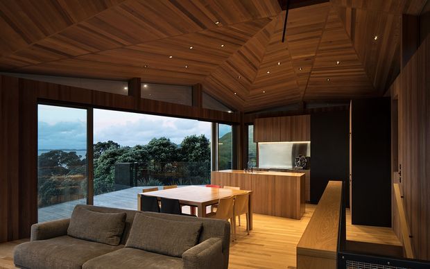 A warm, dark-wood open-plan interior combining a living area, dining space and compact kitchen. The room features extensive timber wall and ceiling lining, a dramatic faceted vaulted ceiling with recessed spotlights, full-height glazing opening to a terrace and wooded coastal landscape, a timber dining table, upholstered sofa, and a streamlined kitchen with timber cabinetry and a pale benchtop.
