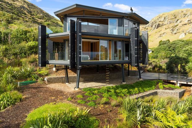A contemporary two-storey house set on a steep, rugged hillside, elevated on slender structural posts. The dark-clad facade features extensive floor-to-ceiling glazing, warm timber-lined upper gable and soffit areas, projecting balconies or terraces, and a low-pitched gabled roof. Native-looking landscaped planting surrounds the building, with a raised walkway connecting to the house.