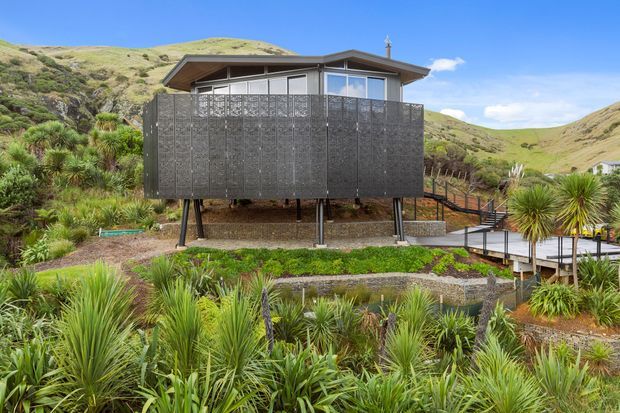 A compact elevated house set into a rugged, landscaped hillside. The dwelling has a low-pitched roof, extensive glazing, and a dark perforated or patterned privacy screen enclosing the raised lower level and deck. Timber and metal stairs connect the building to paths through dense native-looking planting, with rolling dry hills surrounding the site.