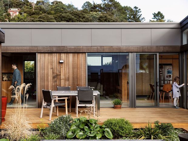 Single-storey contemporary home viewed from a landscaped outdoor deck and garden. The facade combines warm vertical timber cladding with dark-framed floor-to-ceiling sliding glazing, a broad flat roofline, and a covered timber deck furnished for outdoor dining. Interior dining and living spaces are visible through the open and glazed doors, while established vegetation rises behind the home.