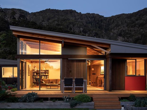 A modern single-storey dwelling set against a densely forested hillside at dusk. The facade combines dark vertical timber cladding, extensive floor-to-ceiling glazing, warm timber soffits and a low-pitched projecting roof. A timber deck with outdoor chairs and steps leads to the warmly lit interior, where a living area, bookshelves and timber-lined spaces are visible.