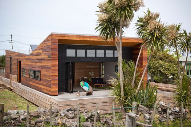 Single-storey contemporary timber-clad house with a strong geometric form and broad covered front opening. The facade combines warm horizontal timber boards with dark window and door framing, while a timber deck and native-looking spiky landscaping surround the entrance. The image is viewed from outside and primarily shows the building exterior.