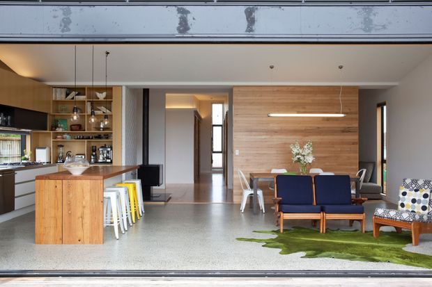 Open-plan contemporary living area viewed through a wide opening, combining a kitchen with timber island and open shelving, a dining zone, and a lounge with mid-century-style seating. The space has polished grey terrazzo flooring, warm timber wall panelling, integrated linear lighting, exposed concrete at the opening, and abundant natural light.