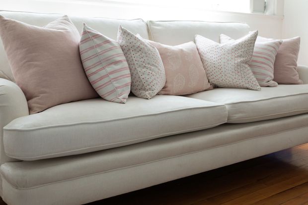 Close-up interior view of a large upholstered sofa with layered blush, white, and pale patterned cushions. The sofa sits on warm-toned timber flooring beside a bright window, creating a soft, comfortable living space with a light neutral palette.