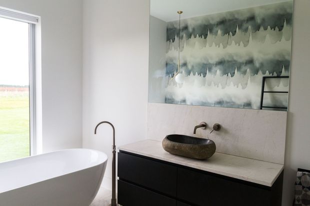 Contemporary bathroom featuring a freestanding white bathtub beside a tall window, a dark floating vanity with a pale stone benchtop, a sculptural vessel basin, wall-mounted bronze-toned tapware, and a large abstract blue-grey and white feature artwork or mural above the vanity.