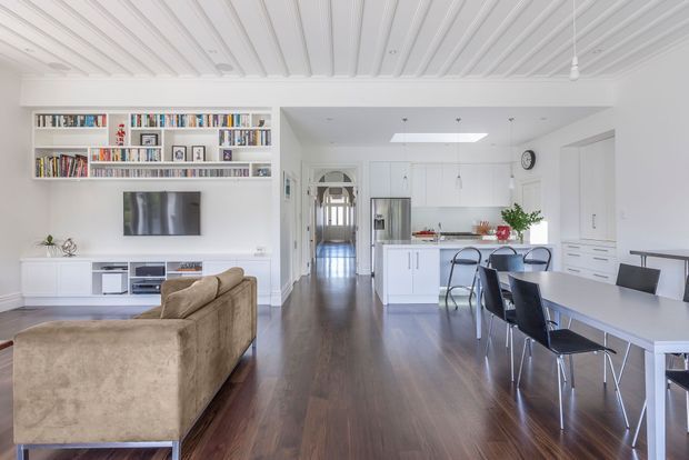 Bright open-plan residential interior combining a living room, dining area and white kitchen. The room features dark timber flooring, white built-in bookcases and media cabinetry, a plush neutral sofa, a long dining table with black chairs, and a distinctive white linear-panel ceiling. A hallway extends beyond the kitchen, creating a spacious connected layout.