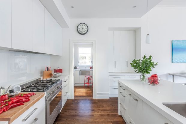 Bright, spacious white kitchen with extensive built-in cabinetry, a large central island, stainless-steel range and refrigerator, glossy white splashback, and dark timber flooring. A small adjoining utility or laundry area is visible through the doorway, while a dining or living area is partially visible at right. The room has a clean, refined contemporary appearance with abundant natural light.