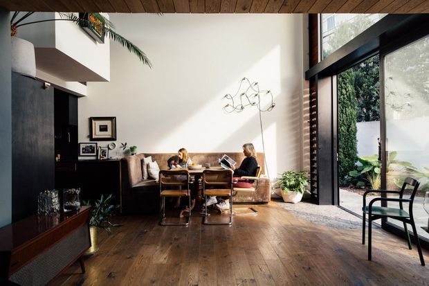 A warm, light-filled mid-century-inspired living and dining space with dark built-in cabinetry, timber flooring, a compact dining table, and a tan sofa along the rear wall. Floor-to-ceiling glazing opens toward a planted courtyard, while exposed timber ceiling boards and black-framed windows add architectural character. A sculptural wall light and potted plants provide understated decorative detail.
