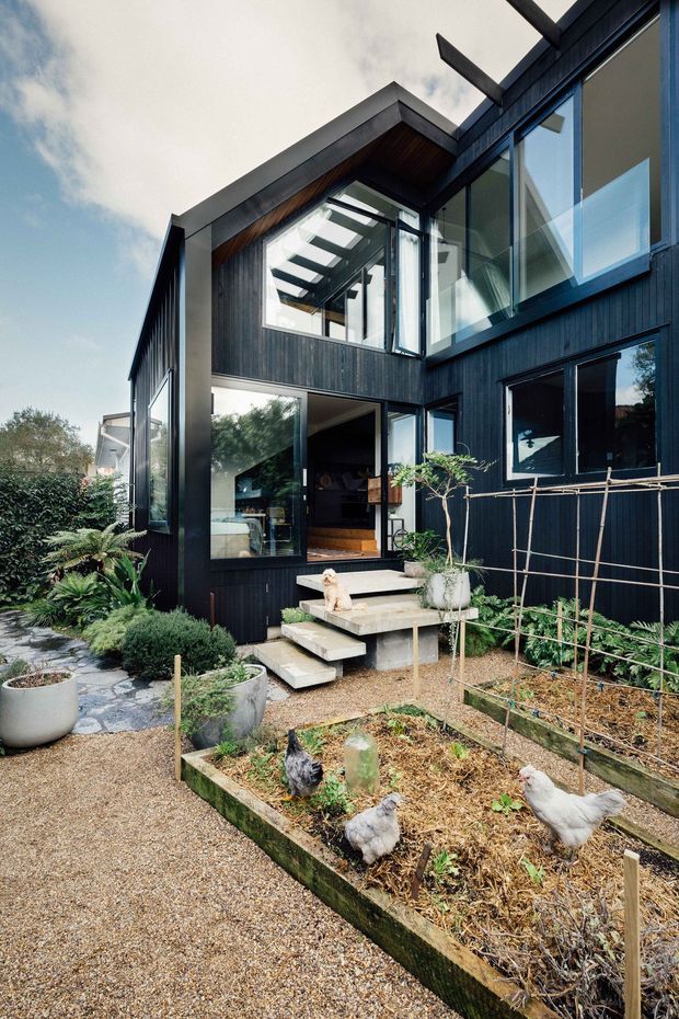Exterior view of a contemporary two-storey home with dark vertical cladding, extensive glazing, projecting roof forms and exposed structural elements. A glazed entry opens onto stepped concrete stairs, with landscaped planting, raised vegetable garden beds and chickens in the foreground.