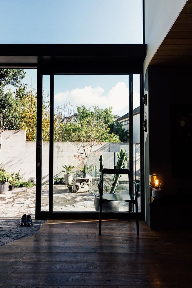 A dark, refined interior looking through full-height black-framed glazed doors into a sunlit paved courtyard. The room has wide dark timber floorboards, a black-framed glass door, a simple black chair, and a small warm table lamp. Outside are potted cacti and other plants against a rendered masonry wall, with neighbouring buildings visible beyond.