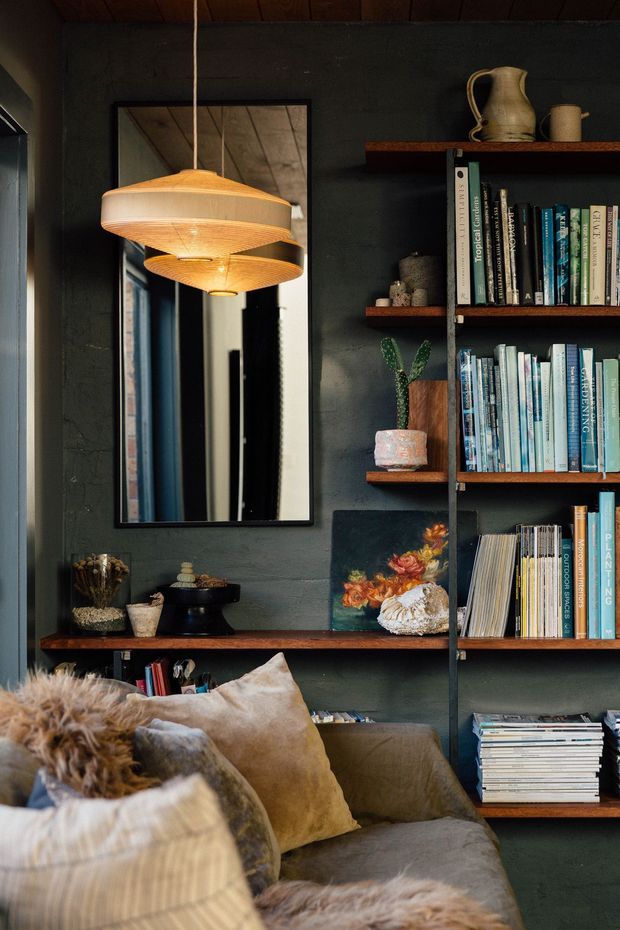 A moody, richly layered living room corner with a deep charcoal textured wall, dark timber ceiling, built-in open shelving filled with books and decorative objects, and a large black-framed mirror. Warm woven pendant lights hang above a plush sofa layered with neutral cushions and faux-fur throws. The styling feels eclectic, cozy, and library-like, with artwork, ceramics, plants, and collected objects adding character.