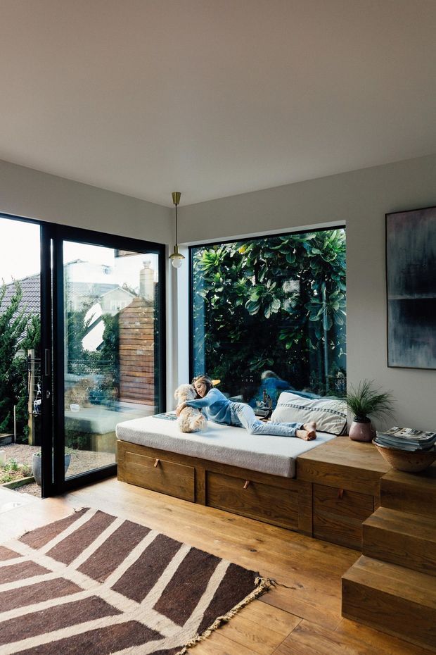 A warm, contemporary living room with floor-to-ceiling black-framed glazing and a glass door opening to a leafy outdoor area. A built-in timber window seat with drawers runs along the window wall, topped with a light upholstered cushion and pillows. A person is reclining on the seat with a small dog. The room has white walls, timber flooring, a patterned geometric rug, a simple pendant light, potted plants, and abstract wall art.