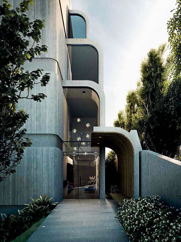 A contemporary multi-storey residential facade viewed from a landscaped entrance path. The building has rounded corners, curved projecting balconies, tall dark-framed glazing, and a distinctive arched entry canopy. The facade appears finished in textured pale blue-grey concrete or render, with a matching curved boundary wall and dense surrounding planting. A glazed entrance reveals a dimly lit interior beyond.