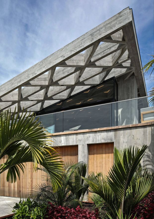 Exterior architectural photograph of a contemporary multi-level building with a dramatic exposed timber gable roof structure, weathered grey concrete walls, dark glazing, and warm timber garage or entry doors. Lush tropical planting frames the foreground, while a balcony or terrace with dark balustrading is visible beneath the roof.