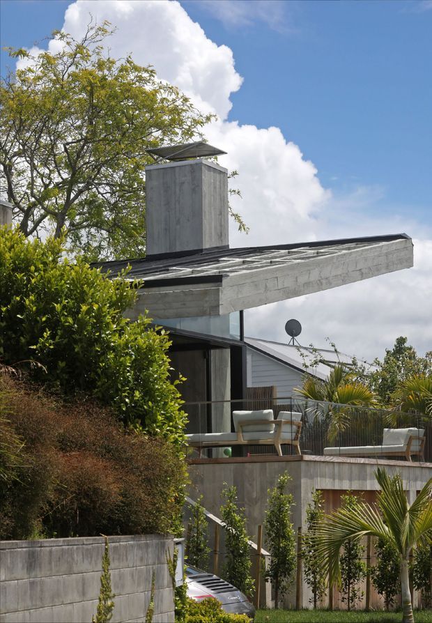 Exterior view of a contemporary multi-level residence on a landscaped sloping site. The building features a flat, projecting concrete roof canopy, a tall concrete chimney or vertical service element, dark metal roof framing, extensive glazing, concrete retaining walls, and elevated outdoor terraces furnished with lounge seating. Palm-like planting and dense garden vegetation surround the structure.