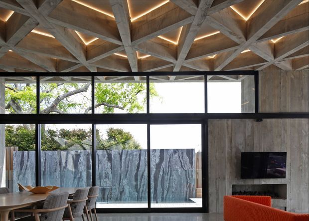 A contemporary living and dining area with expansive black-framed glazing, timber wall cladding, and a dramatic exposed timber roof structure. Warm concealed lighting traces the geometric ceiling beams, while a dining table and chairs occupy the foreground and a bright orange lounge chair sits near a built-in fireplace and television. A leafy tree and sculptural exterior screening are visible through the windows.