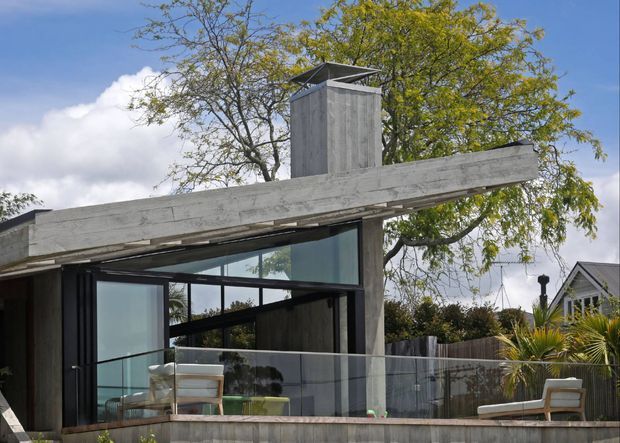 Exterior view of a modern single-storey pavilion or extension with a dramatic exposed concrete beam and vertical concrete chimney-like element. The structure has extensive dark-framed glazing, a transparent glass balustrade, and a small outdoor seating area. Mature trees, fencing, and a neighbouring gabled house are visible in the background.