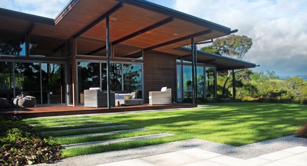 Contemporary single-storey residence with broad timber-lined roof overhangs, dark slender framing and extensive floor-to-ceiling glazing. The house opens onto a manicured lawn via broad stone stepping slabs and shallow landscaped steps, with outdoor seating visible beneath the covered terrace. The setting appears leafy and coastal or subtropical, with a bright, open indoor-outdoor connection.