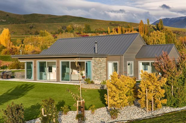 Single-storey contemporary home set in a landscaped rural or mountain setting, with grey metal gabled roofs, muted horizontal cladding, natural stacked-stone wall accents, and large glazed doors and windows. The property has a manicured lawn, gravel garden beds, deciduous planting, and expansive views toward rolling hills and mountains.