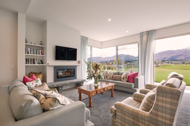 Bright, comfortably furnished living room with a corner of floor-to-ceiling glazing overlooking a green valley and mountains. The room features pale upholstered sofas and plaid armchairs, a timber coffee table, built-in open shelving, a wall-mounted television, and a contemporary fireplace with a stone-look surround. Soft curtains, cushions, and floral arrangements add a relaxed, homely feel.