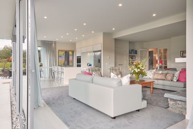 Bright, open-plan contemporary living area with a large light-grey sectional sofa, pale rug, patterned accent cushions, timber coffee table, and a stone-clad fireplace or hearth. The space opens directly to a modern white kitchen with integrated cabinetry and stainless-steel appliances, and features extensive glazing, sheer curtains, recessed ceiling lights, and a view toward an outdoor terrace.