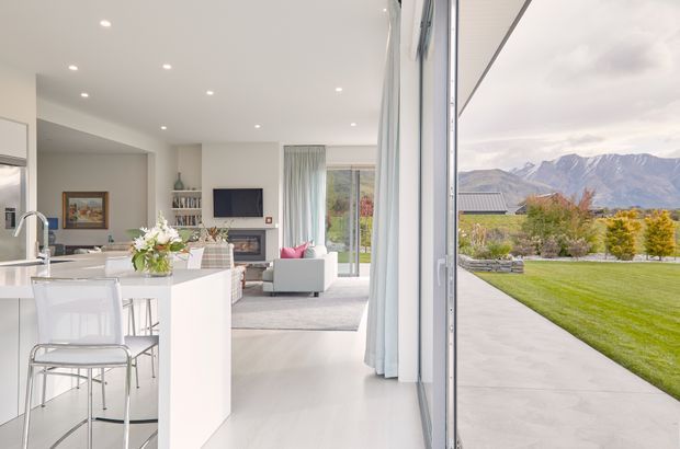 Bright open-plan contemporary interior with a white kitchen island and bar stools in the foreground, opening into a minimalist living room with a fireplace, television, shelving and pale furnishings. Full-height glazed sliding doors connect the room to a broad lawn and paved terrace, with dramatic mountains visible beyond the property.
