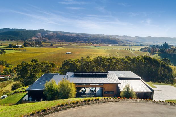 Aerial exterior view of a low-profile contemporary building set on a landscaped, elevated site. The building has multiple intersecting metal-clad roof forms, dark-framed glazing, and warm masonry walls, with expansive agricultural fields, wooded hills, and a broad rural valley beyond.