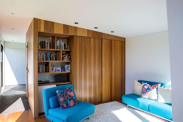 Bright contemporary living room featuring extensive floor-to-ceiling timber built-in cabinetry with integrated shelving, books, and concealed storage. A vivid turquoise armchair and sofa with colourful patterned cushions sit on a light textured rug, with strong daylight entering from the left and a hallway visible beyond.