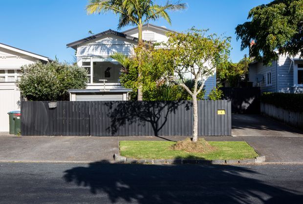 Street-facing view of a compact residential property with a light weatherboard facade, enclosed front porch, garage, dark vertical timber fence, driveway, and a small grass verge. Tropical and leafy garden planting surrounds the house under clear daylight.