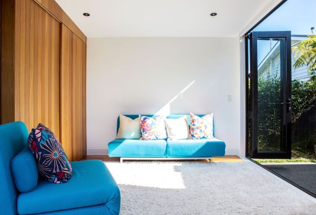 Bright contemporary living room with vivid turquoise upholstered seating, a large pale rug, full-height warm timber built-in wall panelling, and colourful patterned cushions. The room opens directly through black-framed glazed doors to a small planted outdoor area, with strong natural sunlight and a clean white ceiling featuring recessed downlights.
