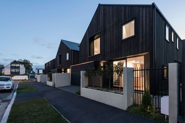 Exterior architectural photograph of a contemporary multi-unit residential development at dusk. Several narrow, two-storey homes feature steep gabled roof forms, dark vertical timber-like cladding, black-framed windows, recessed warm lighting, and small landscaped front courtyards enclosed by low rendered walls and black metal fencing. A paved shared driveway and neighbouring houses are visible.