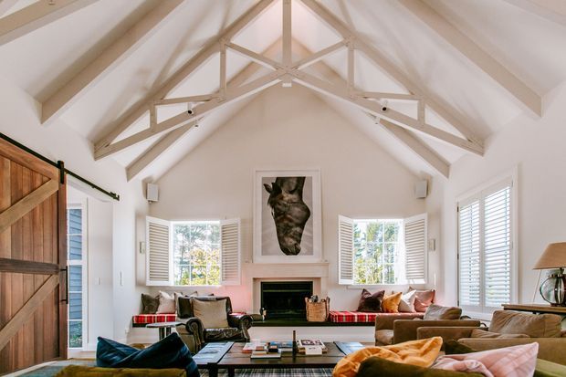 Bright, spacious living room with a vaulted white ceiling and exposed decorative trusses, white walls, shuttered windows, a central fireplace, and layered seating around a low coffee table. Timber barn-style doors and warm upholstered furnishings add a relaxed rustic character.