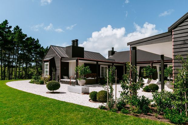 Exterior view of a contemporary single-storey home arranged around a landscaped courtyard and outdoor living area. The dark horizontal cladding, pitched metal roofs, covered patio, white gravel paving, clipped shrubs, and mature trees create a refined modern garden setting.