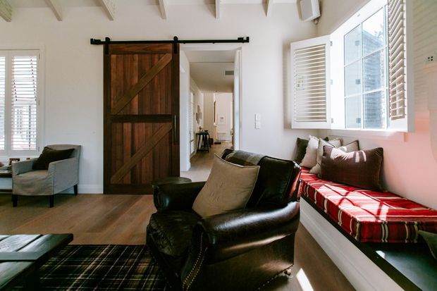 Bright, inviting living room with white walls, exposed ceiling beams, timber flooring, plantation shutters, and a rustic dark timber barn-style sliding door. A black leather armchair and sofa seating face a low coffee table, while a built-in window seat with red plaid upholstery adds colour and warmth.