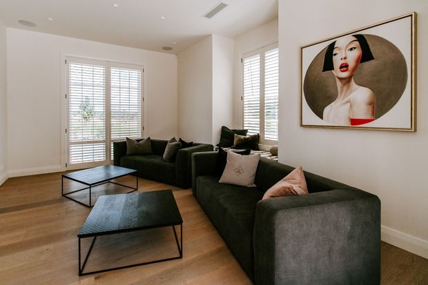 Bright contemporary living room with white walls, warm timber flooring, large shuttered windows and a dark green sectional sofa arranged around two low black metal coffee tables. A framed portrait artwork adds a bold focal point to the otherwise neutral interior.