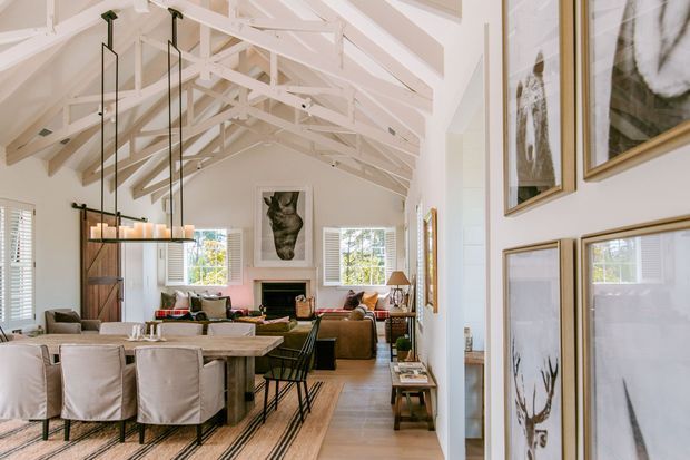 Bright open-plan living and dining room with a high vaulted ceiling, exposed white-painted timber trusses and beams, white walls, timber flooring, and extensive framed artwork. A large dining table with upholstered chairs occupies the foreground, while a seating area, fireplace, and decorative shelving are visible beyond. Contemporary pendant lighting and abundant windows create an airy, refined interior.