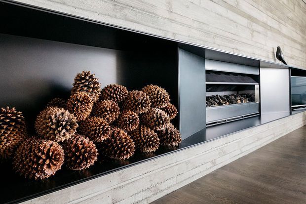 Close-up of a contemporary built-in display wall featuring black recessed niches, pale horizontal timber wall panelling, and integrated shelving. Decorative pine cones fill the left niche, while the central shelf contains stacked firewood. A dark timber-look floor and minimal black detailing complete the refined interior.