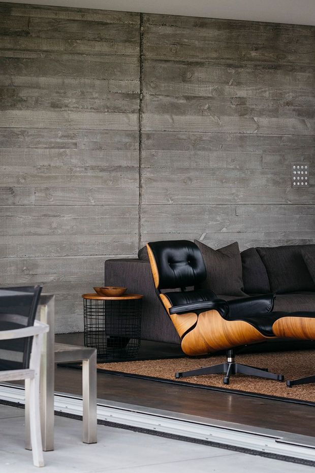 A moody, industrial-style living room featuring a dark charcoal sofa, black leather and warm wood lounge chair, black wire side table, and textured grey concrete wall panels. A dining table and chairs are partially visible in the foreground, separated from the living area by a large sliding glazed opening or track.