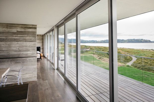 A long, bright contemporary interior with full-height glazed sliding doors opening onto a timber deck and glass balustrade. The space features a concrete or concrete-look feature wall, dark timber flooring, and views across landscaped coastal or waterfront terrain. A partial dining or living area is visible at the left.