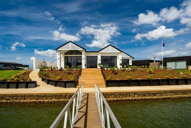 Front exterior view of a low-rise contemporary gable-roofed building beside a body of water, approached by a narrow pedestrian bridge and broad timber steps. The facade combines white rendered walls, dark-framed glazing, twin gabled roof forms, landscaped raised garden beds, and a waterfront retaining edge under a bright blue sky.