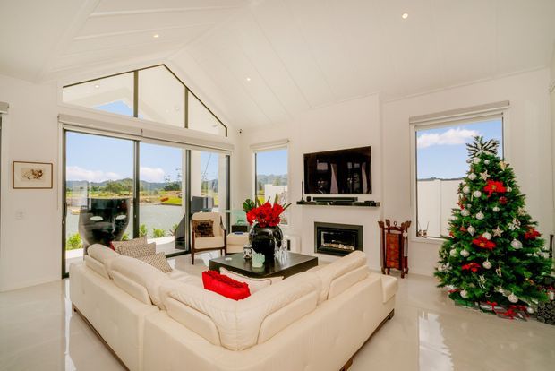 Bright, spacious contemporary living room with a large cream sectional sofa arranged around a dark coffee table, wall-mounted television, and built-in fireplace. Floor-to-ceiling glazing and a raised triangular clerestory-style window provide abundant natural light and views outside. The room has glossy light flooring, white walls and ceiling, and festive Christmas decorations including a decorated tree.