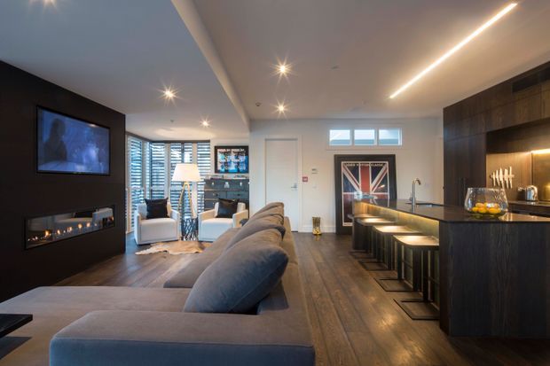 Open-plan contemporary living and kitchen interior with a large grey sectional sofa facing a wall-mounted television and linear fireplace. The kitchen features dark timber cabinetry, a dark stone island with integrated seating, pendant lighting, and warm under-cabinet illumination. Timber flooring, recessed spotlights, and timber venetian blinds create a refined, moody atmosphere.