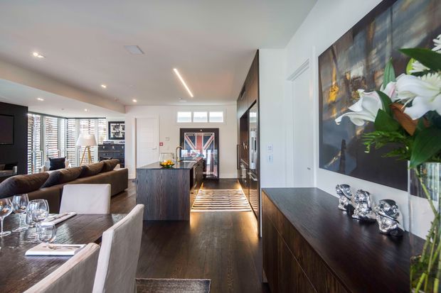 Open-plan contemporary interior combining a dining area, living room, and kitchen. Dark timber flooring and cabinetry contrast with light walls, upholstered dining chairs, a large sectional sofa, linear recessed lighting, and a prominent floral artwork above a sideboard. The kitchen island visually anchors the centre of the space, while glazing and a glazed entry door bring in natural light.