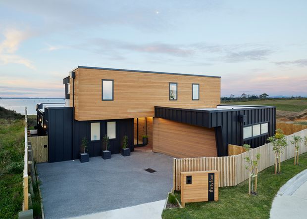 Contemporary two-storey house with a rectilinear form, warm horizontal timber cladding, dark metal-clad lower volumes, and a projecting upper level. The home includes a covered garage or entry volume, paved driveway, timber fencing, and sparse landscaping. It is photographed from the street or driveway with open coastal-looking surroundings and a wide sky backdrop.