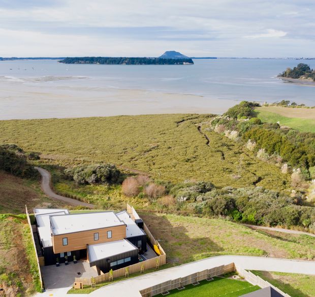 Aerial view of a modern flat-roofed residence set within a landscaped coastal or estuarine site. The two-storey home has light timber-toned cladding, dark trim, paved outdoor areas and a fenced lawn, with expansive tidal water, vegetated shoreline and a low wooded island in the background.