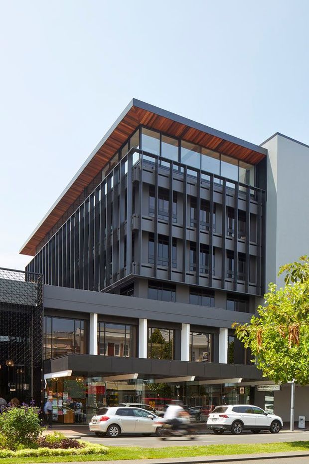 Street-level photograph of a contemporary multi-storey commercial or mixed-use building. The facade combines dark vertical metal fins and extensive glazing with a projecting pitched roof featuring warm timber soffit lining. The lower level contains glazed retail or commercial frontage, while the upper levels have balconies or screened openings. Landscaping, pedestrians, and parked vehicles occupy the foreground.