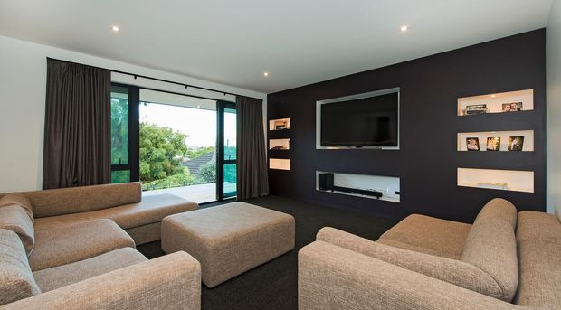 A contemporary living room centred on a dark charcoal feature wall with a wall-mounted television, recessed display niches and a built-in media console. Multiple large neutral upholstered sofas and an ottoman surround the room, while full-height glazed doors/windows with dark curtains open the space to leafy outdoor views. Recessed ceiling lights and a dark carpet create a comfortable, home-cinema-like atmosphere.